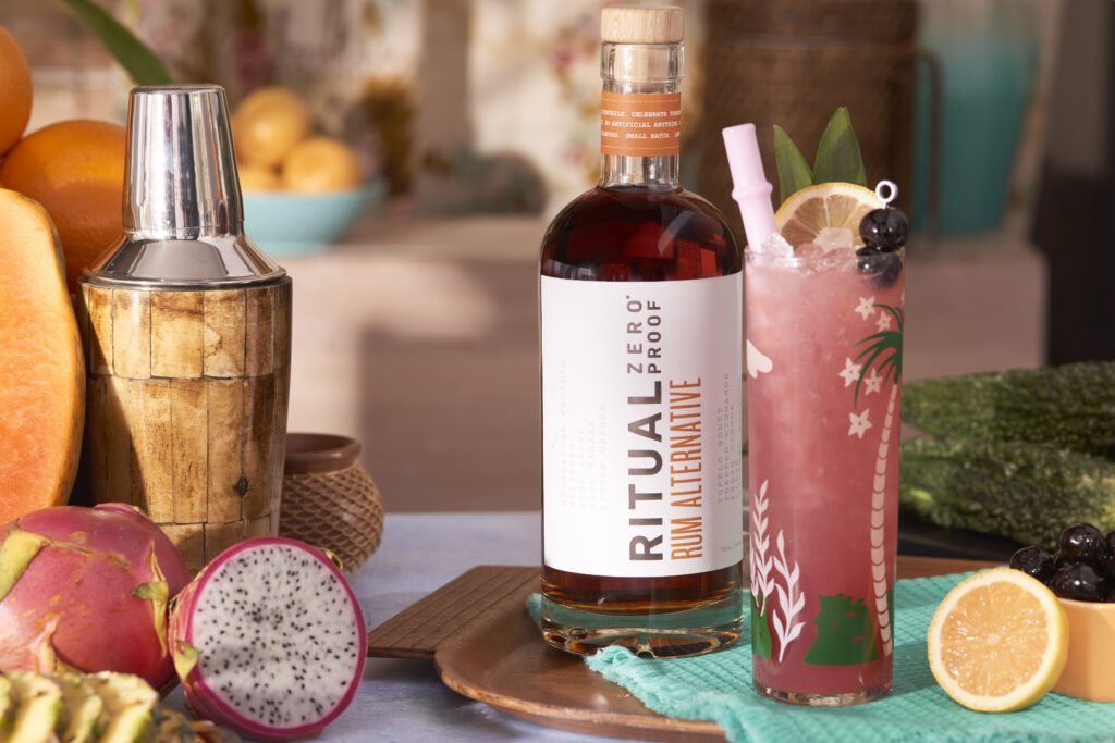 A tropical pink cocktail sitting on a wooden tray surrounded by fruits and a shaker.